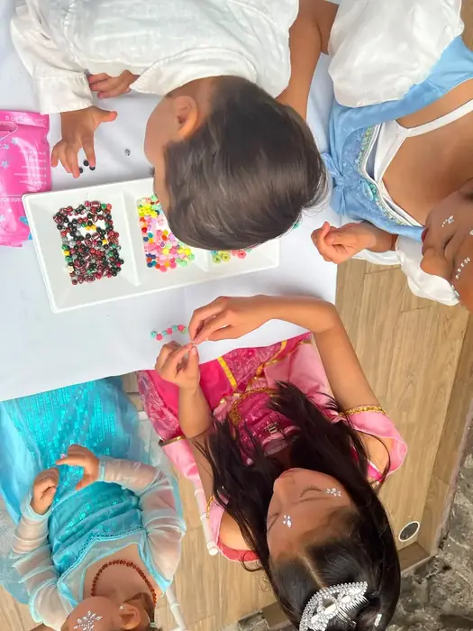 Niñas vestidas de princesas armando pulseras de cuentas de colores en una mesa de manualidades