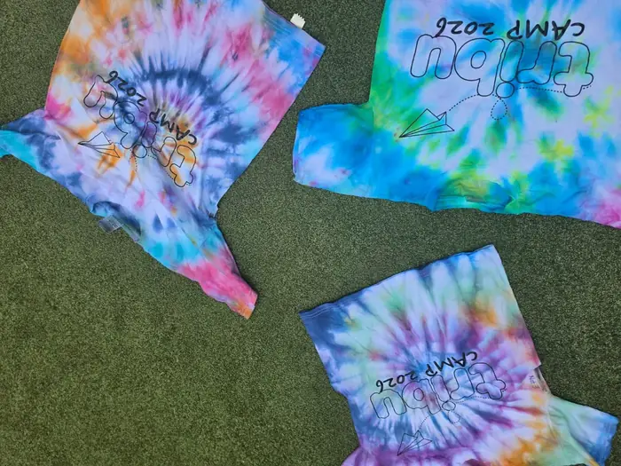 Playeras teñidas a mano con la técnica tie-dye y el logo de Tribu Camp 2026, tendidas sobre el pasto